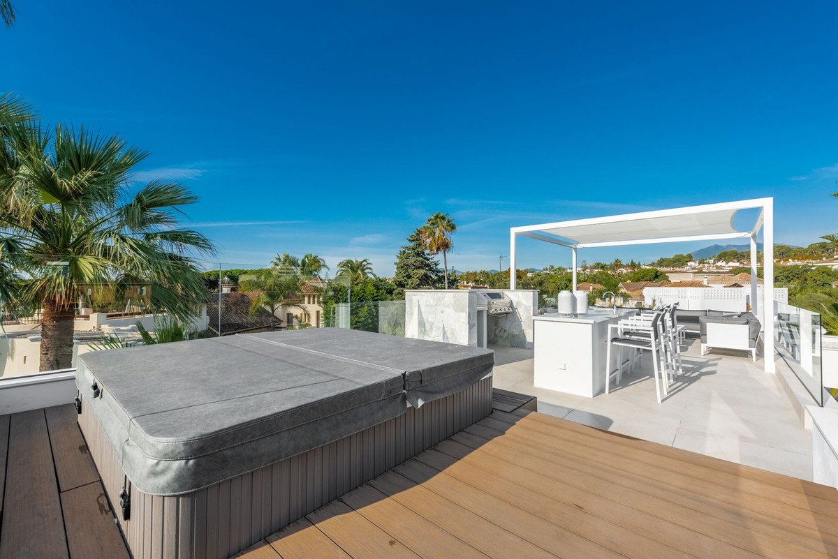 A spacious rooftop terrace featuring a covered hot tub, outdoor kitchen with a grill, bar seating, and lounge area with a distant view.
