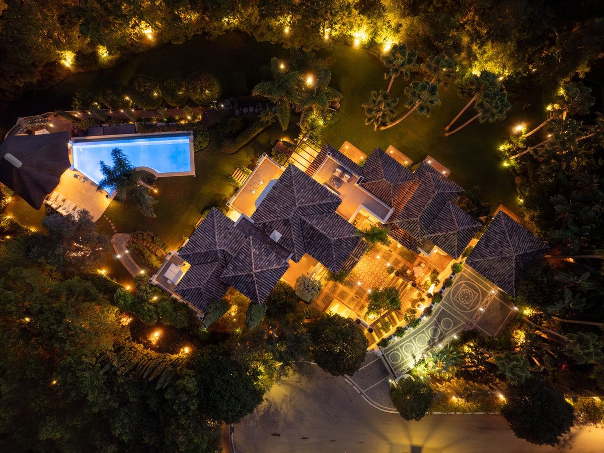 Aerial view of a luxury villa at night, featuring a swimming pool, illuminated gardens, and tiled rooftops, surrounded by lush trees.