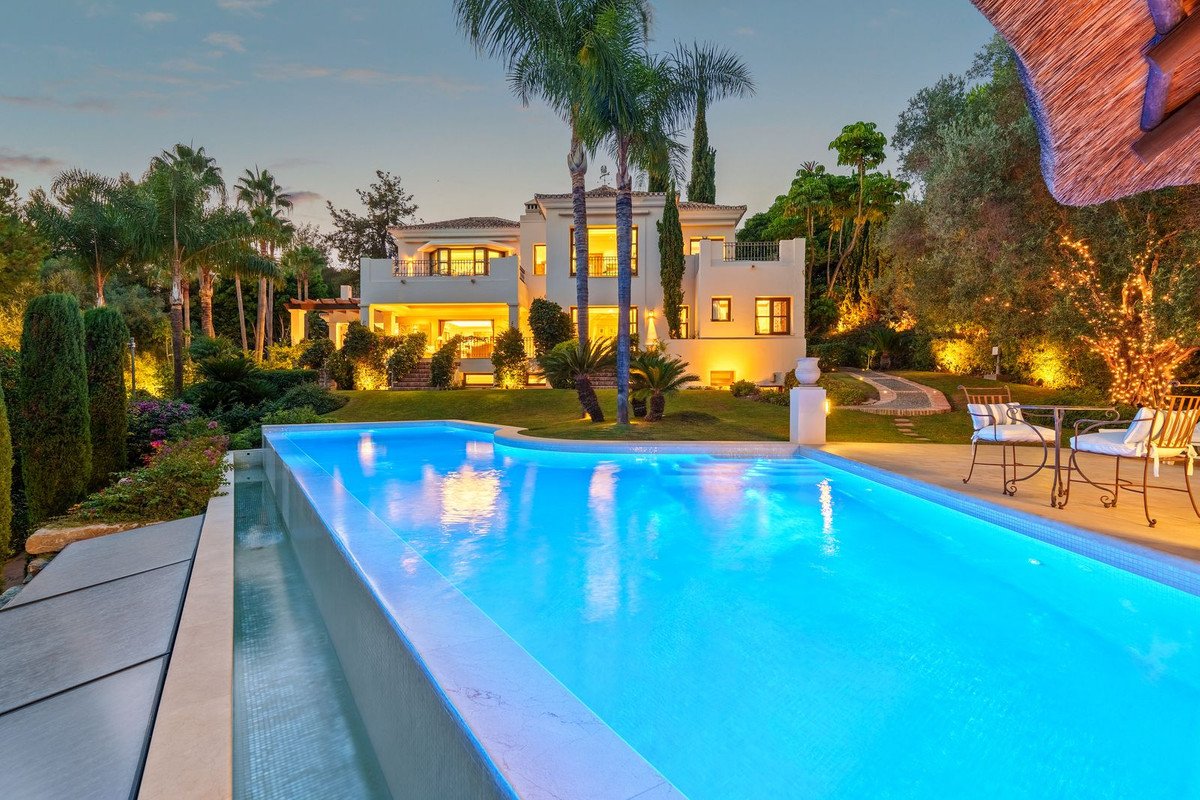 Luxurious villa with an infinity pool and outdoor seating area at dusk, surrounded by tropical gardens and illuminated trees.