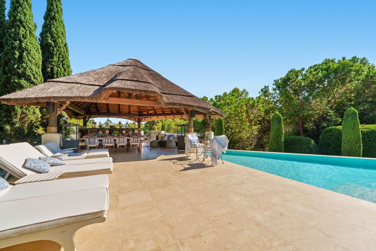 An outdoor thatched-roof dining area with a long table, pool, and lounge chairs, surrounded by trees and a blue sky.