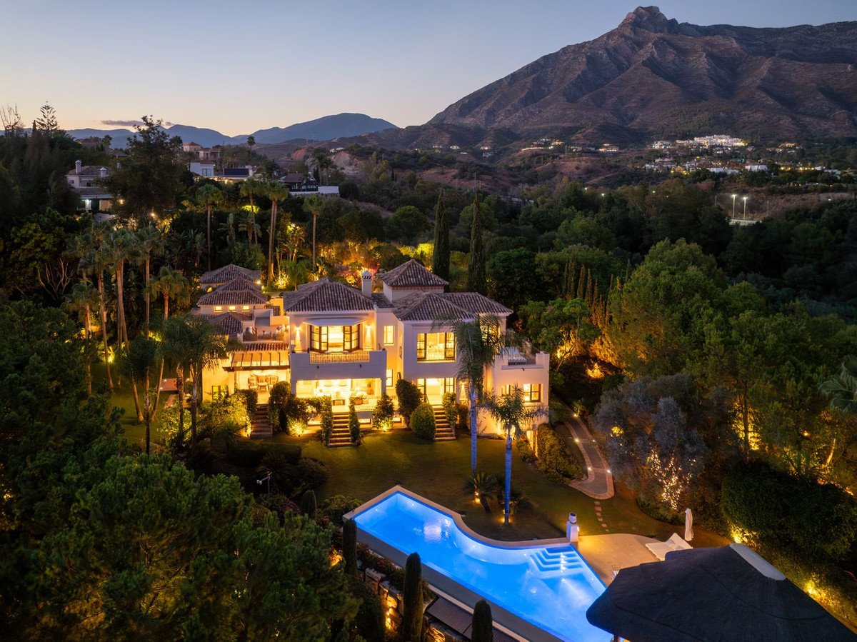 Luxurious villa with illuminated pool and expansive gardens set against a backdrop of mountains at dusk.
