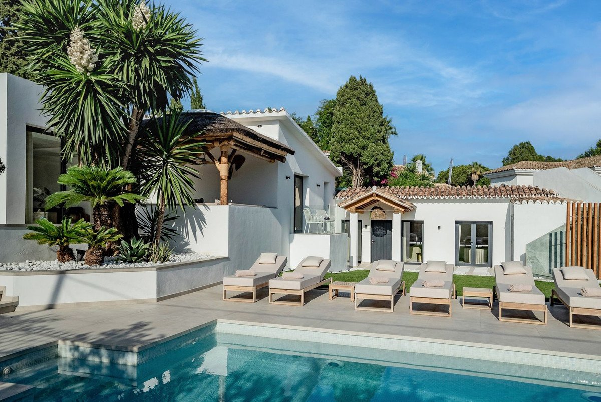 A modern villa with a swimming pool, lounge chairs, and lush tropical landscaping on a sunny day.