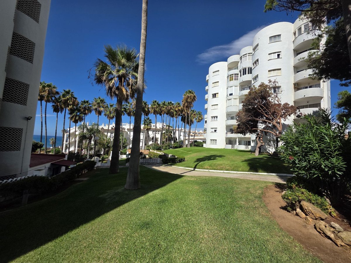 White apartment buildings surrounded by lush green grass and tall palm trees, with a distant view of the sea under a clear blue sky.