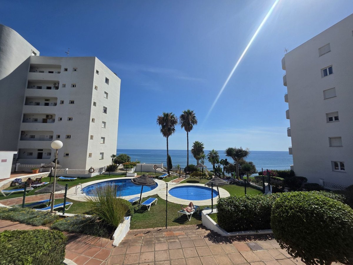 Apartment complex with two swimming pools, lush greenery, palm trees, and a beautiful sea view under a clear blue sky.