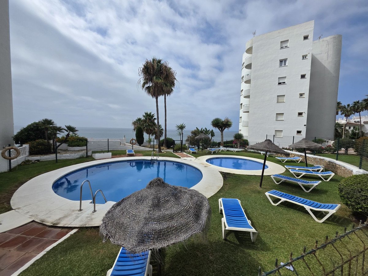 Two swimming pools with loungers and umbrellas, overlooking the sea, with palm trees and a modern building.