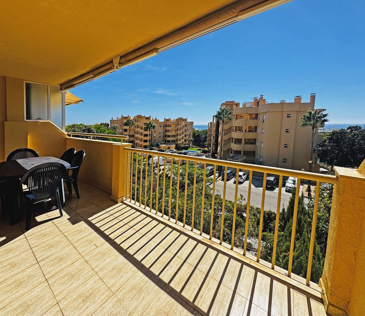 A balcony with a table and chairs overlooks apartment buildings and a distant view of the sea on a sunny day.
