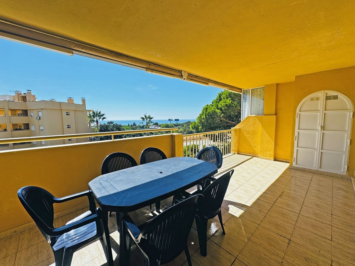 A sunny balcony with a dining table and chairs, offering a distant sea view and overlooking buildings and trees.