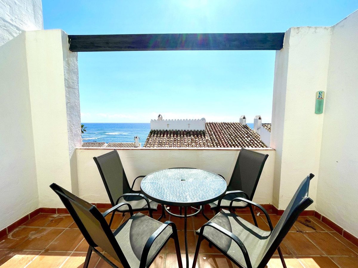 A balcony with a glass table and four chairs offers a view of the sea and rooftops under a clear blue sky.