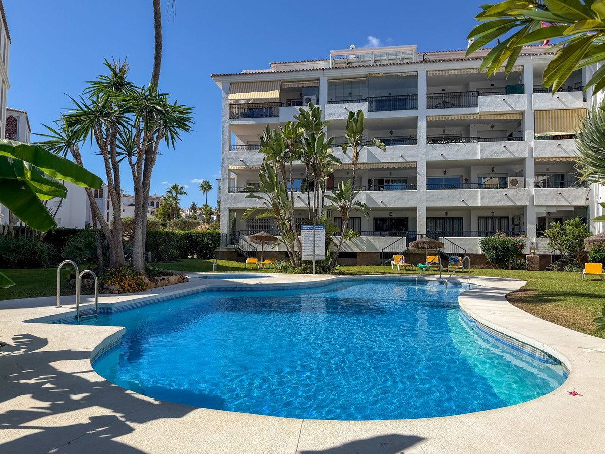 A bright blue swimming pool in front of a white apartment building with balconies and awnings, surrounded by palm trees and lush greenery.