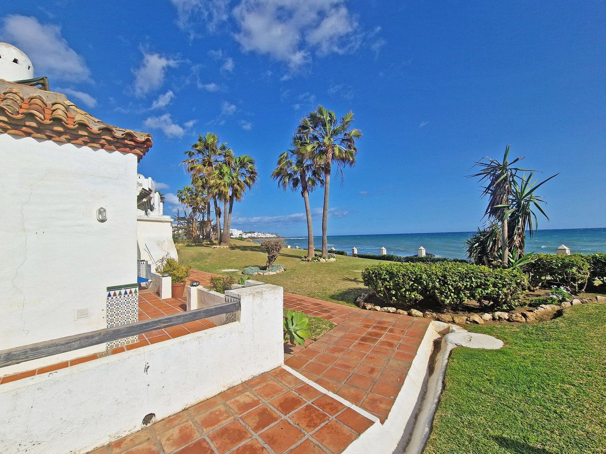 A white building with a tiled terrace overlooks a grassy garden with palm trees and the blue sea beyond.