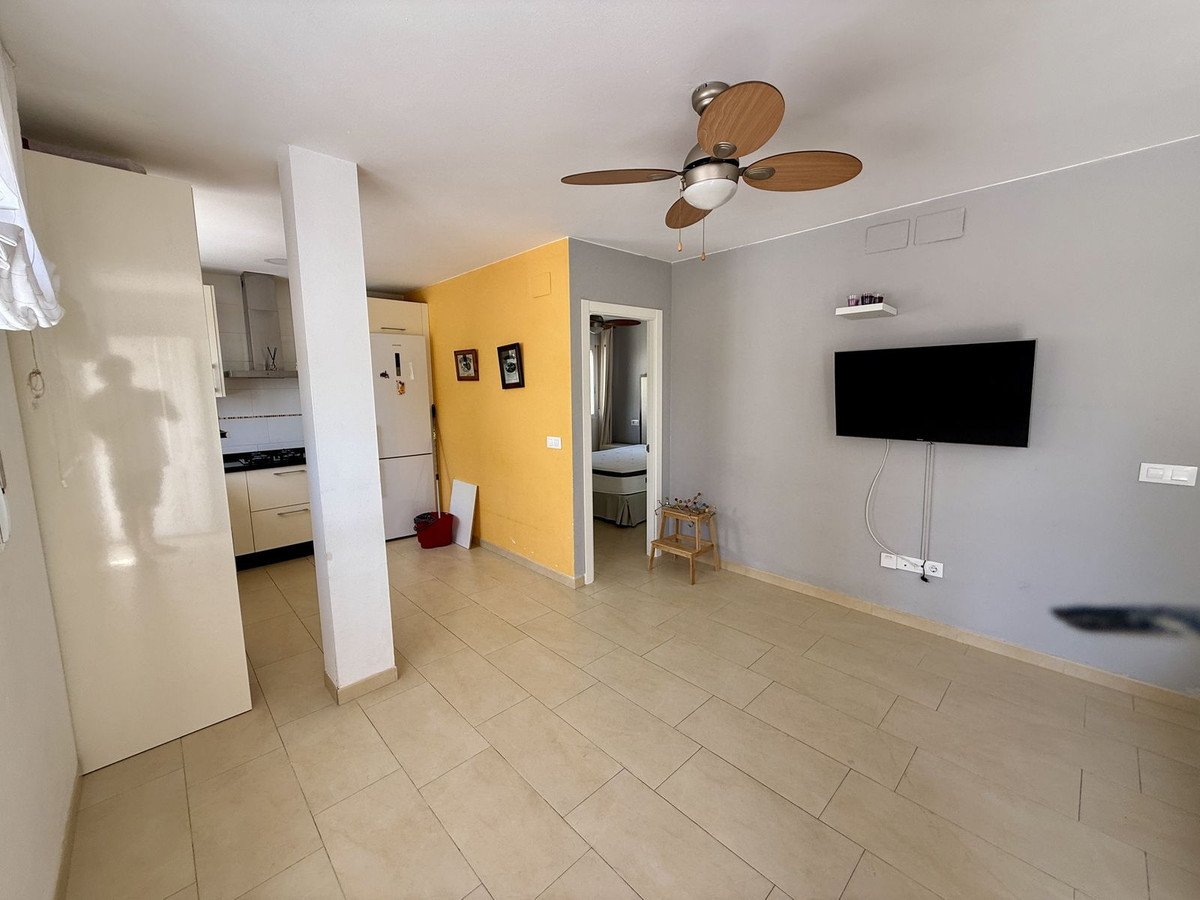 A modern living area with an open-plan kitchen, featuring a wall-mounted TV, ceiling fan, and tiled floors.