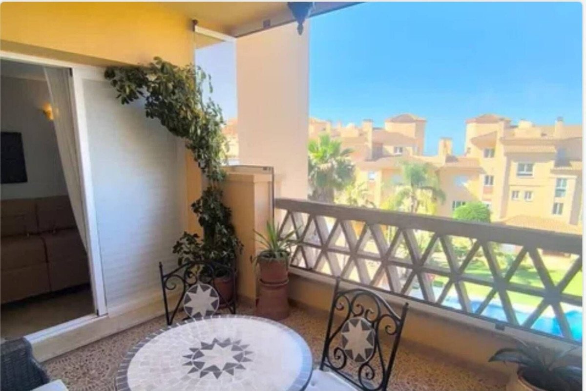 A balcony with a mosaic table and chairs, offering a view of buildings and a swimming pool on a sunny day.