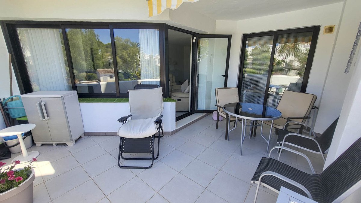 Spacious balcony with outdoor seating, a table, and chairs, overlooking a bright residential area with greenery.