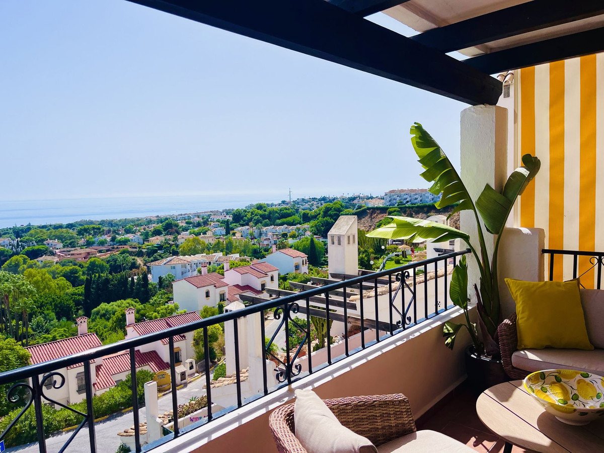 A balcony with furniture offers a stunning sea view over a residential area with green trees and white houses.