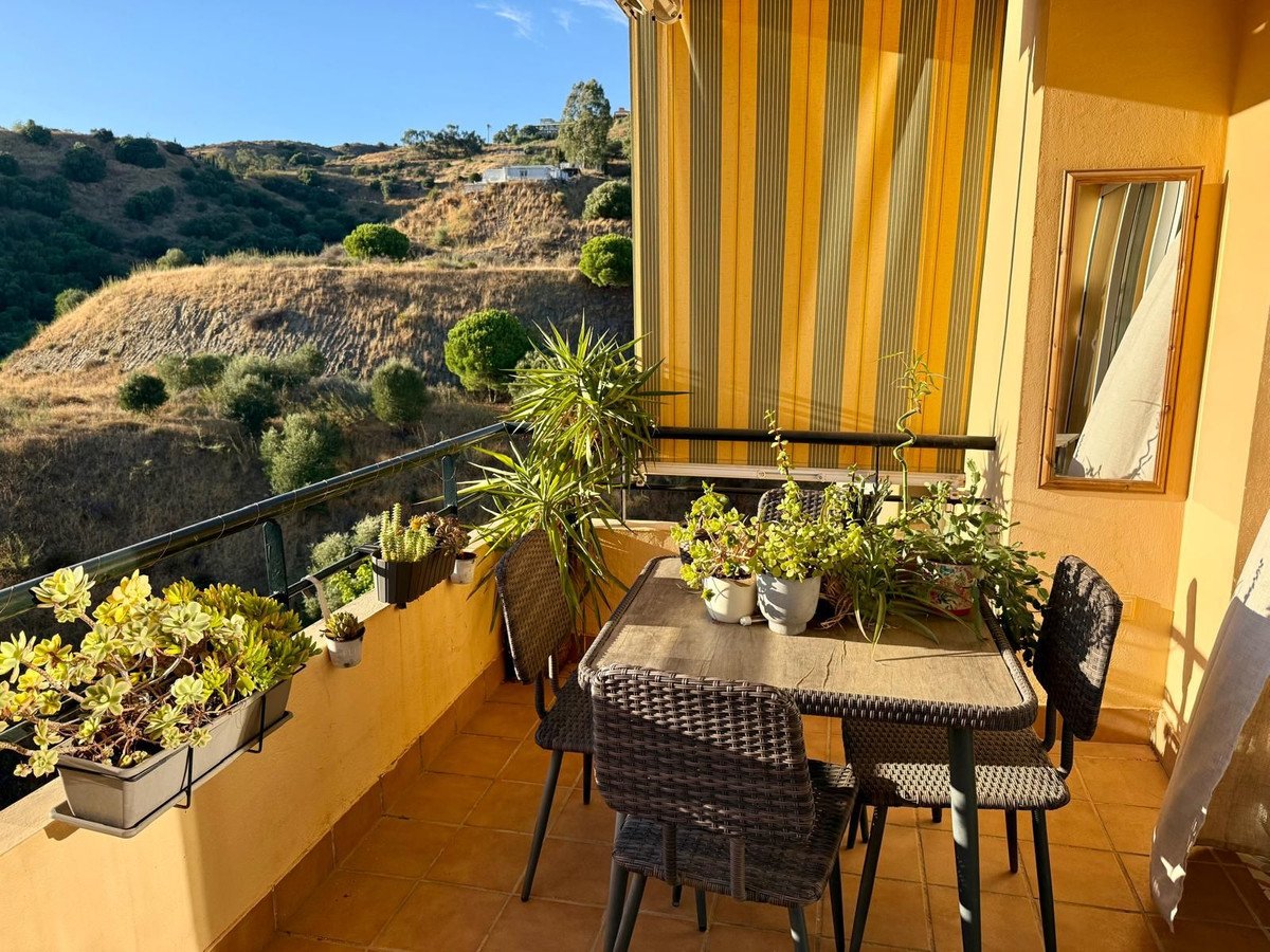 A sunny balcony with a table, chairs, and potted plants, offering a view of rolling hills.