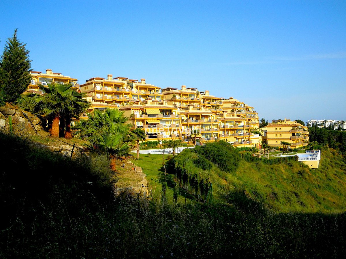 Apartment complex with yellow facades and balconies on a lush green hillside under a clear blue sky.