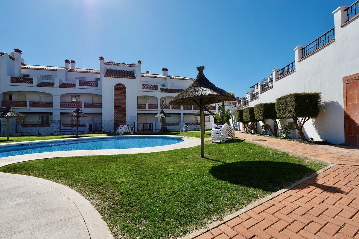 Apartment complex with a swimming pool, grass, sun loungers, and straw umbrellas on a sunny day.