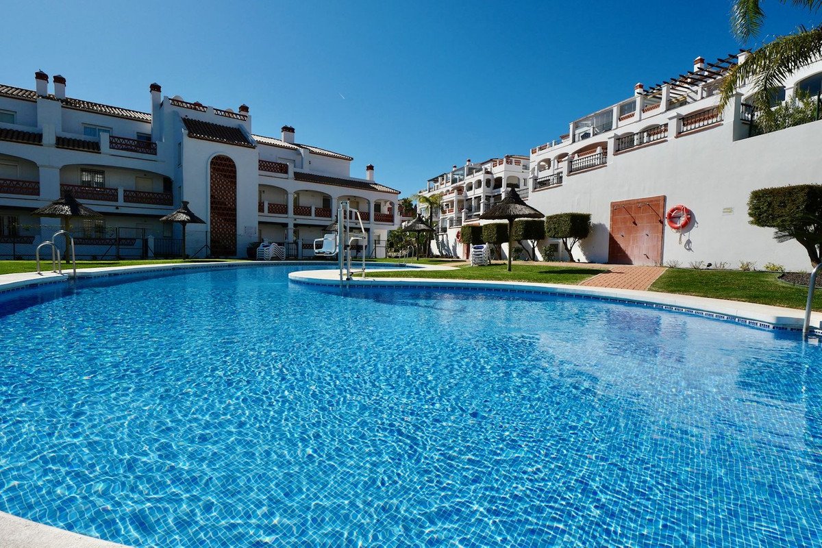 A large, sparkling blue swimming pool with lounge chairs and umbrellas, surrounded by white apartment buildings under a clear blue sky.