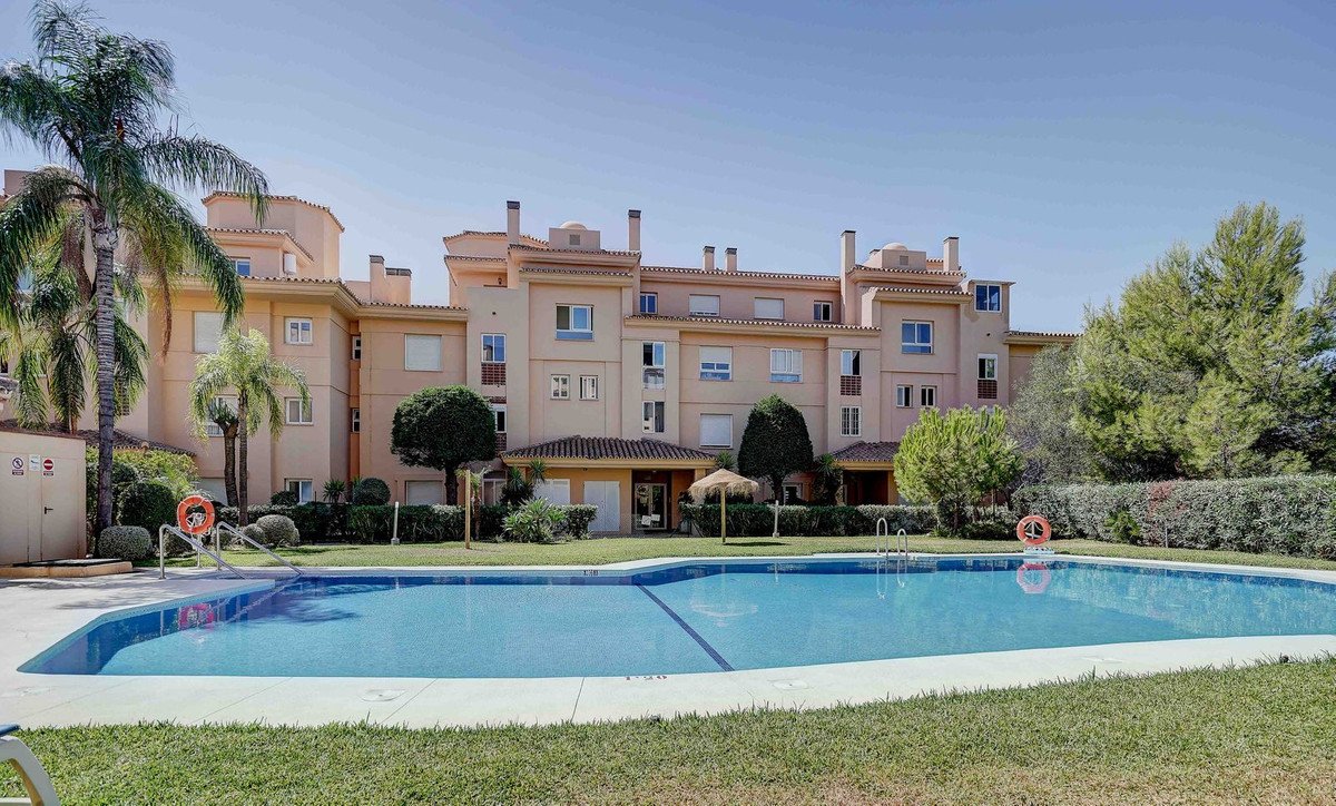 Exterior of a beige apartment building with a large blue swimming pool and lush green grass in front.