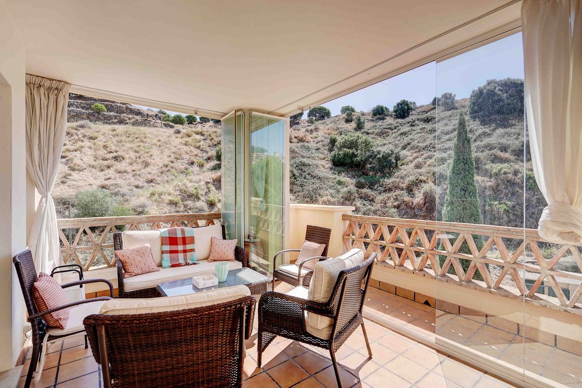 A covered terrace with rattan furniture, offering a scenic view of a dry, hilly landscape and surrounding trees.