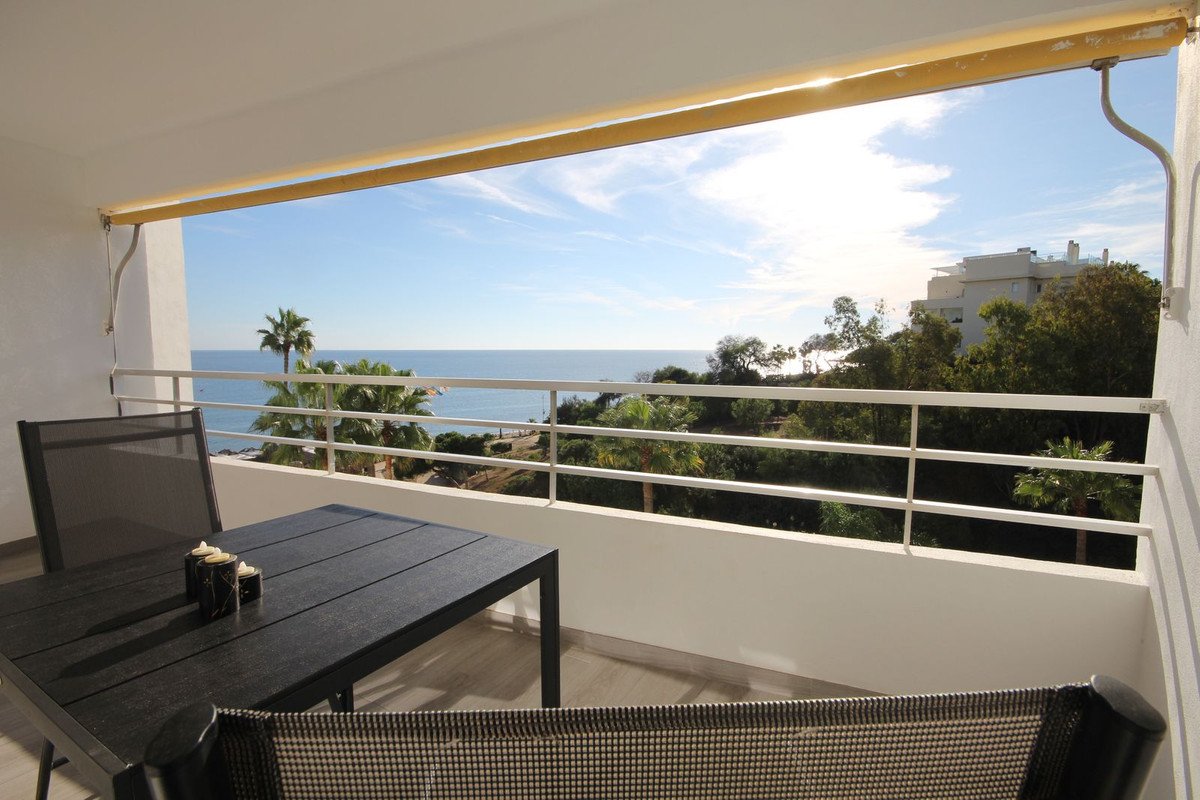 Balcony with a table and chairs overlooking the sea, with palm trees and a building in the background on a sunny day.