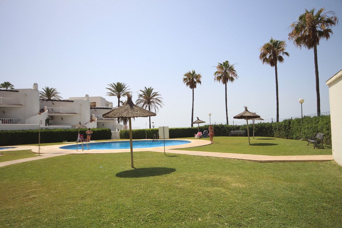 A sunlit communal garden with a swimming pool, palm trees, and a distant sea view.
