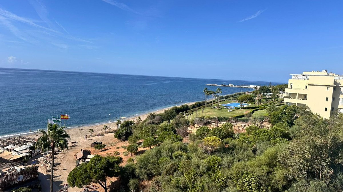 View from apartment balcony overlooking a beach, sea, swimming pool, and resort buildings.