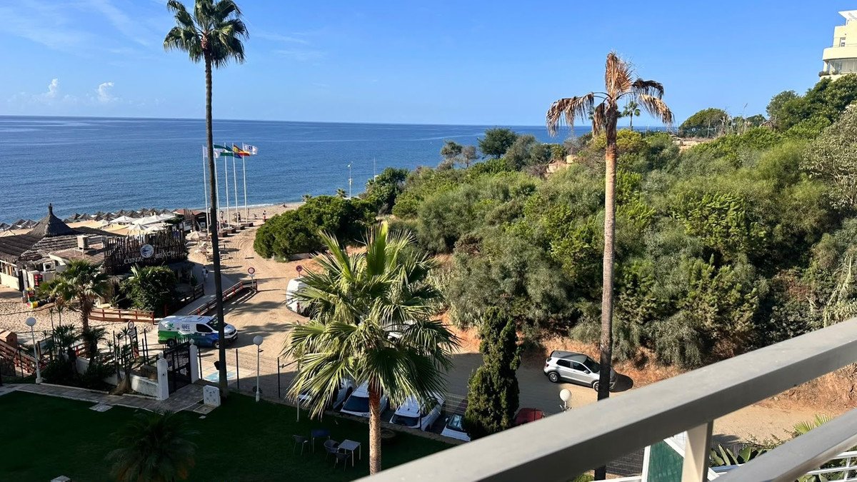 A scenic view from a balcony overlooking the sea, with palm trees and a beachside restaurant below.