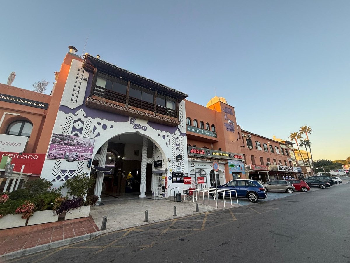 Commercial building with an archway, shops, and parked cars on a sunny day.