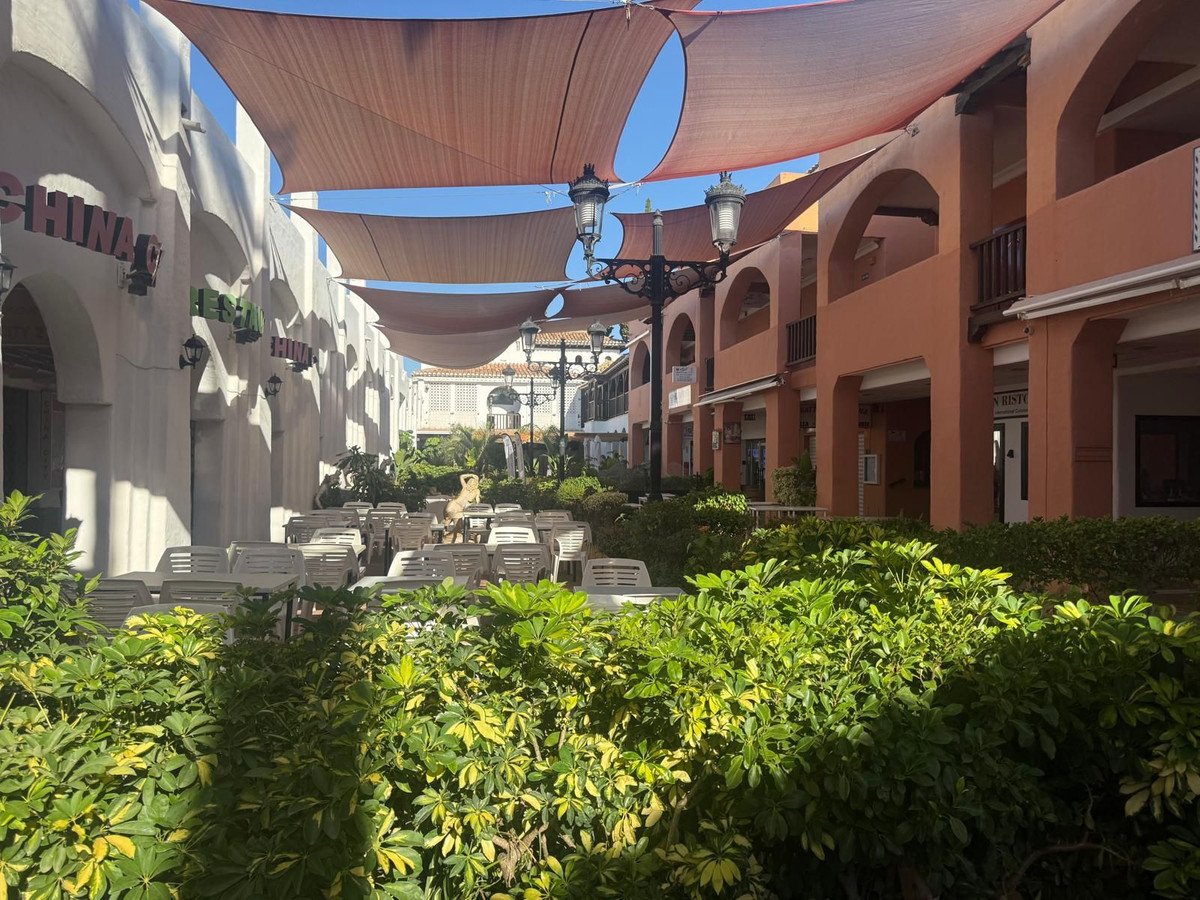 An outdoor cafe seating area with tables and chairs covered by sun shades, surrounded by lush green plants and buildings.