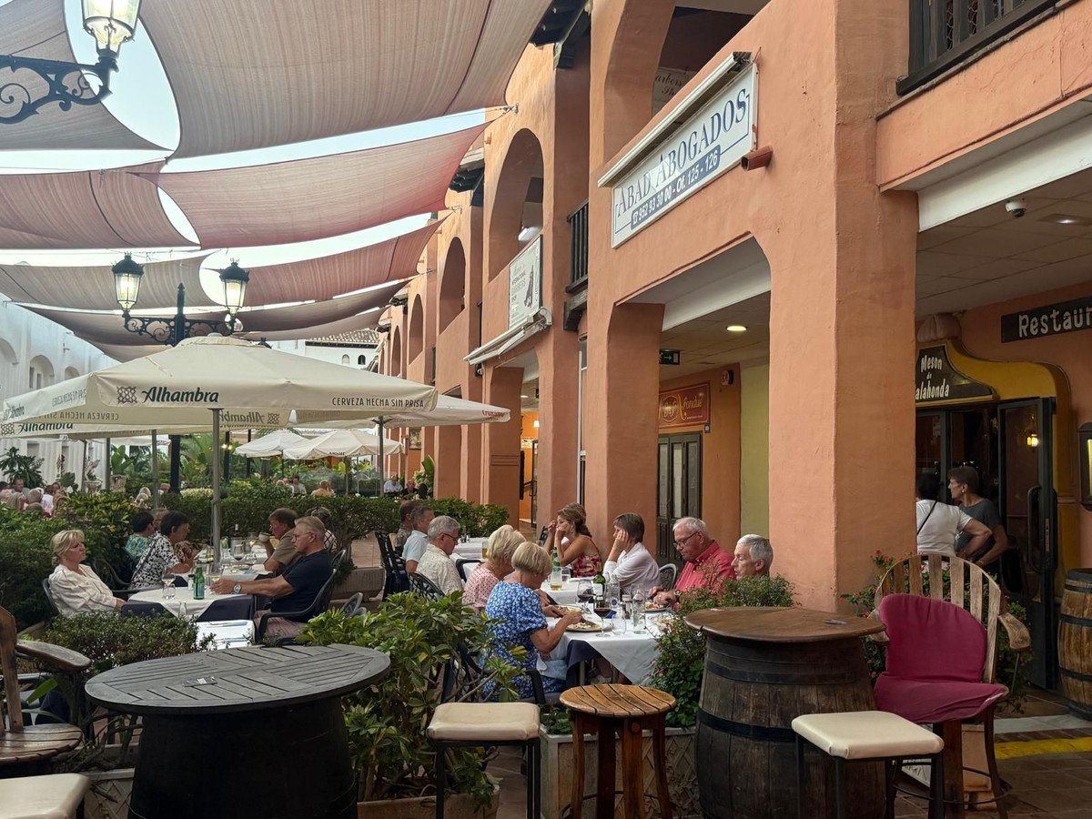People dining at tables on a restaurant patio under awnings, with arched walkways and greenery.