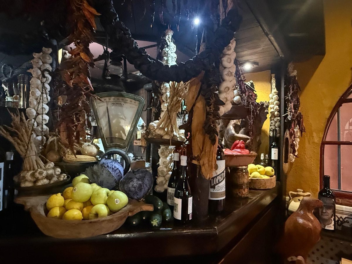 A rustic display of dried peppers, garlic, lemons, apples, and other produce hangs above a bar with bottles and a vintage scale.