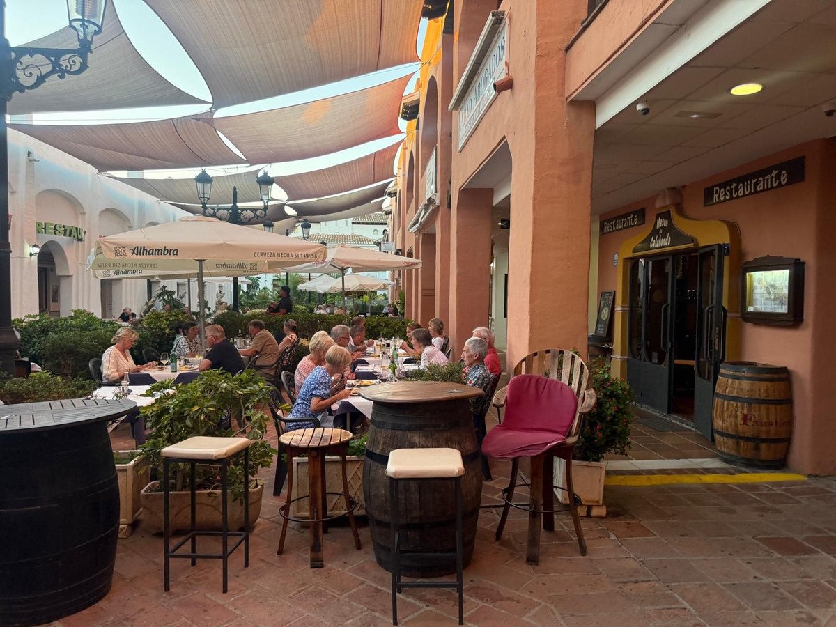 People dining at tables on an outdoor restaurant patio under awnings, with plants and barrels.