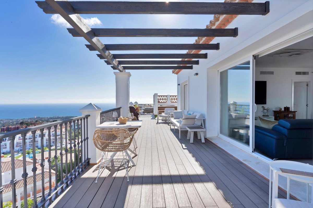 Spacious terrace with outdoor furniture, overlooking the sea and coastal town under a pergola and sunny sky.