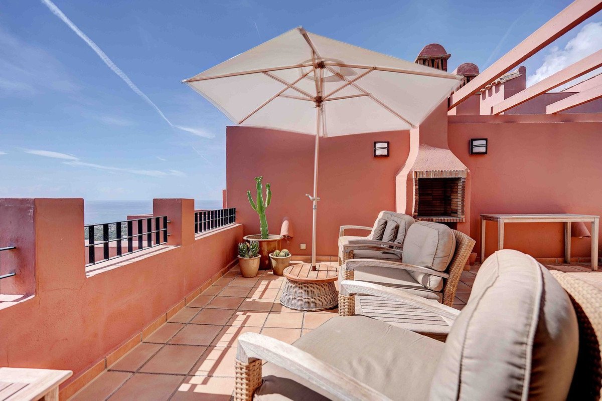 Terrace with seating, umbrella, and grill, offering a sea view under a blue sky.