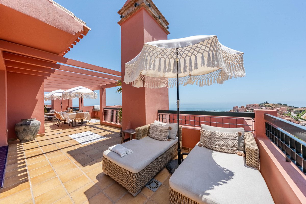 A terrace with two sun loungers and umbrellas, overlooking the sea and a coastal town under a clear blue sky.