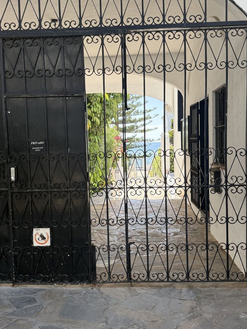Wrought iron gate opens to a path with a view of the sea and trees in the distance.