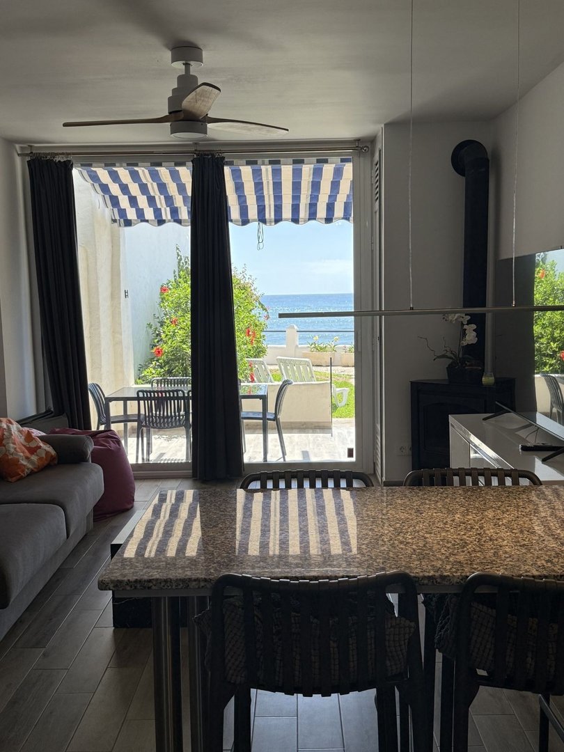 Interior view of a living and dining area with a sea view and outdoor terrace accessible through glass doors.