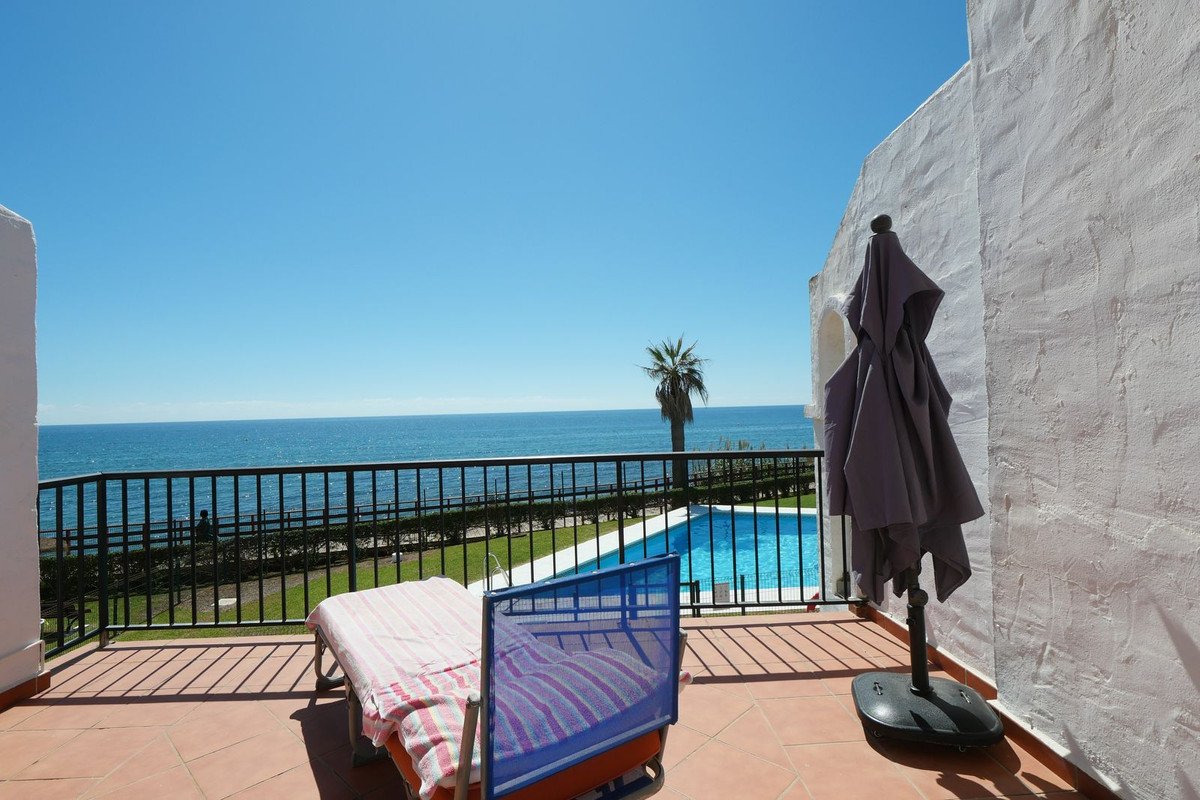 A sunny balcony with a lounger and umbrella overlooks the sea, pool, and a palm tree.
