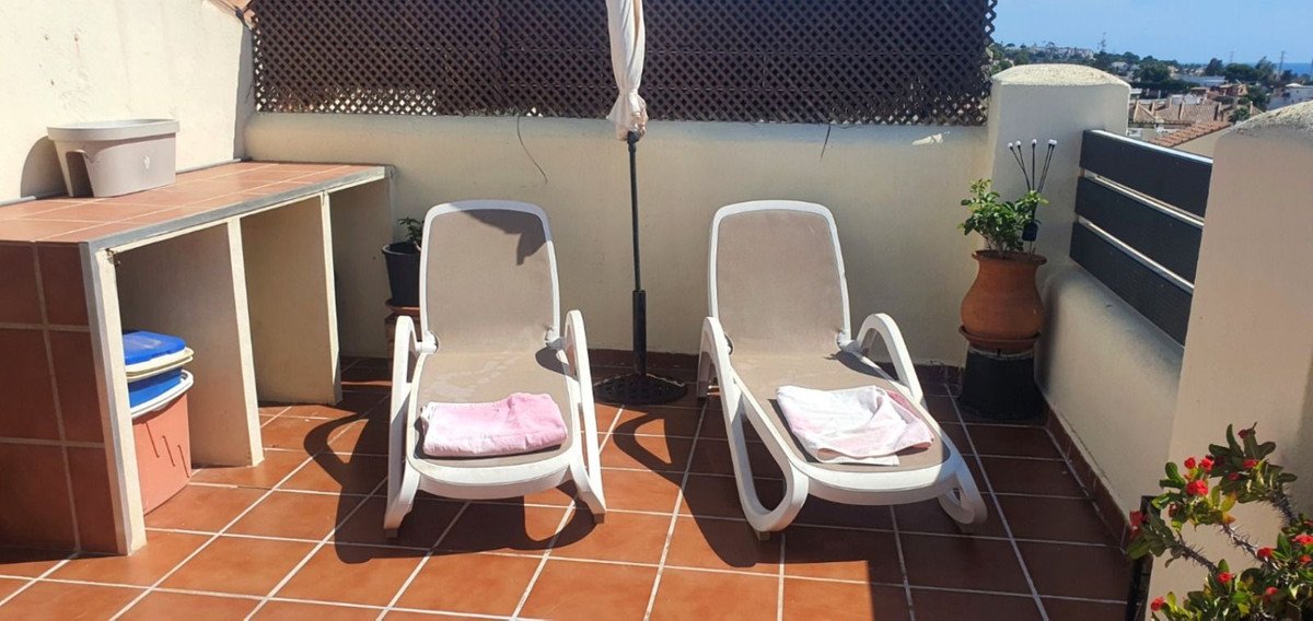 Two lounge chairs with towels on a tiled terrace with an outdoor kitchenette and a distant view of rooftops and the sea.