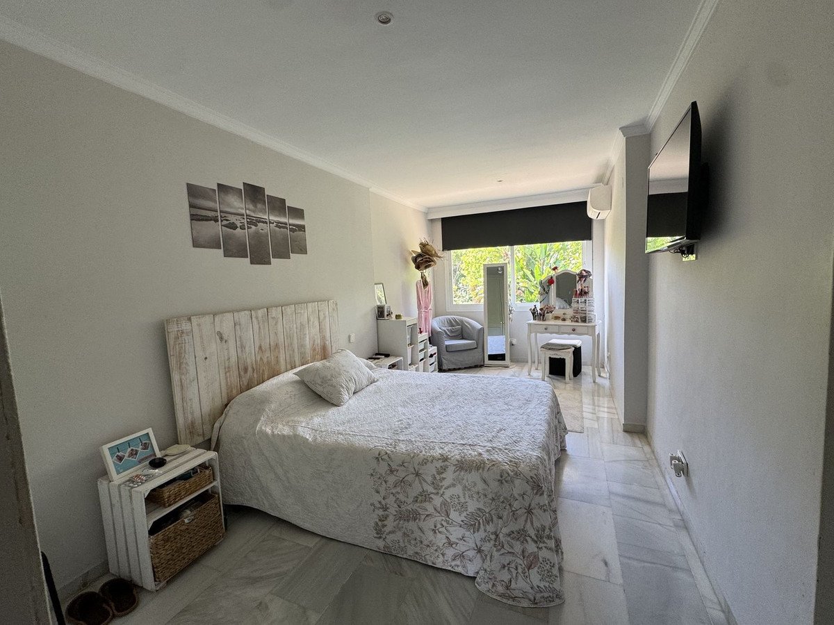 A bright bedroom with a wooden headboard bed, white furniture, and a view of greenery through a large window.