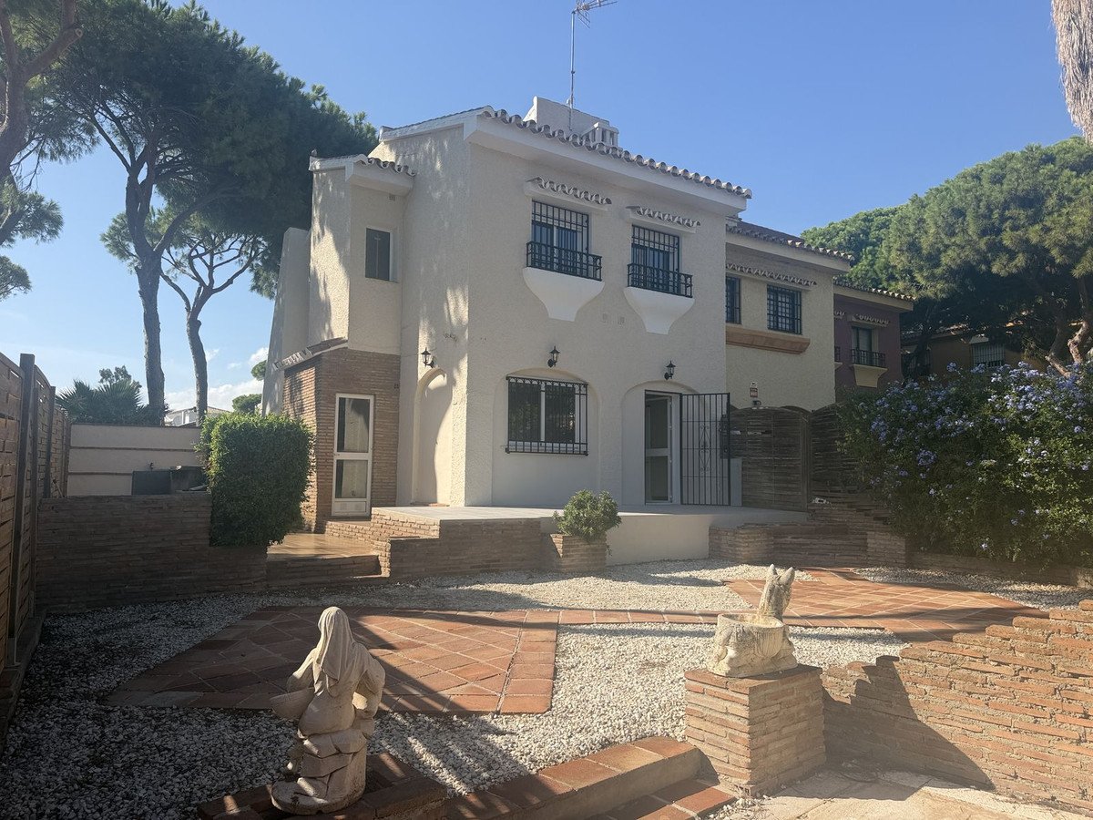 Exterior view of a villa with a terracotta-tiled patio, garden, and lush greenery on a sunny day.