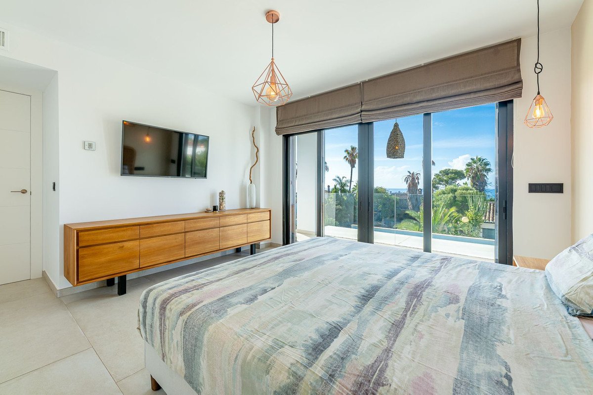 Bedroom with a large sliding glass door leading to a balcony with a sea view and palm trees. Includes a TV and modern furniture.