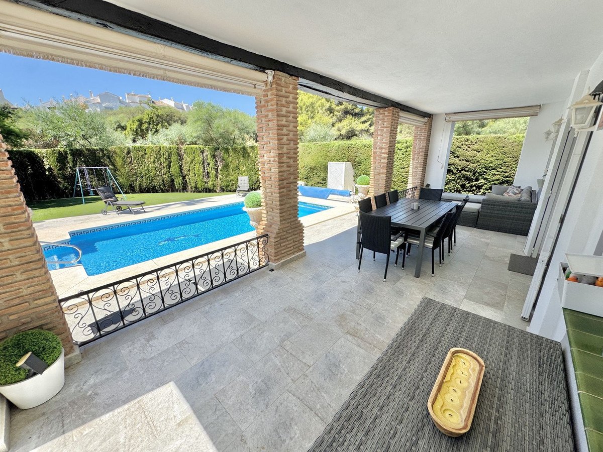 Covered patio with dining table and sofa, overlooking a swimming pool and garden with a swing set.