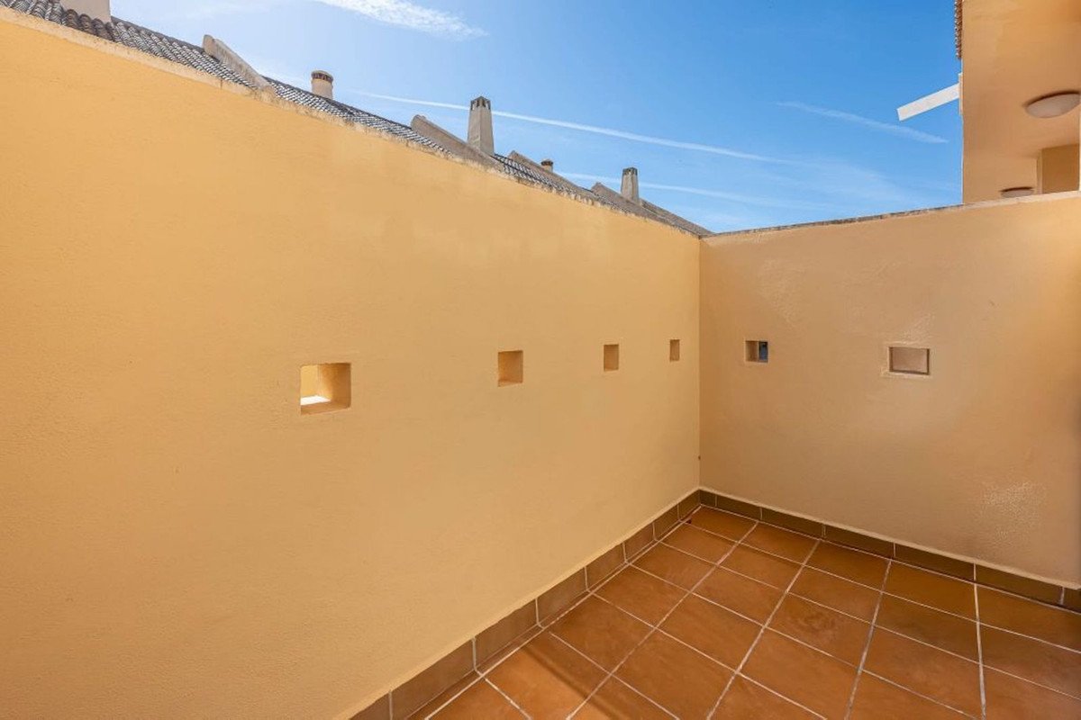 A corner of a terrace with terracotta tiled floor, beige walls, and square openings, under a blue sky.
