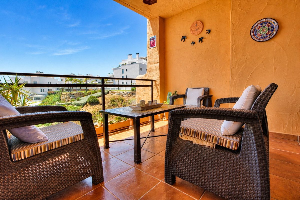 Terrace with wicker chairs and table overlooking a sunny landscape with modern buildings and palm trees.