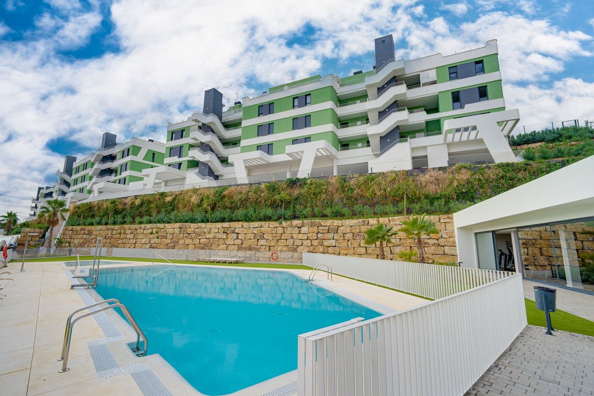 Modern apartment complex with a swimming pool and gym, set against a hillside under a cloudy sky.