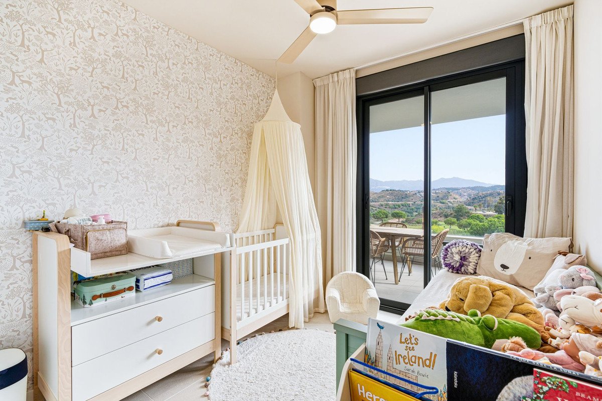 A nursery room with a crib, changing table, stuffed animals, and a balcony overlooking rolling hills and mountains.