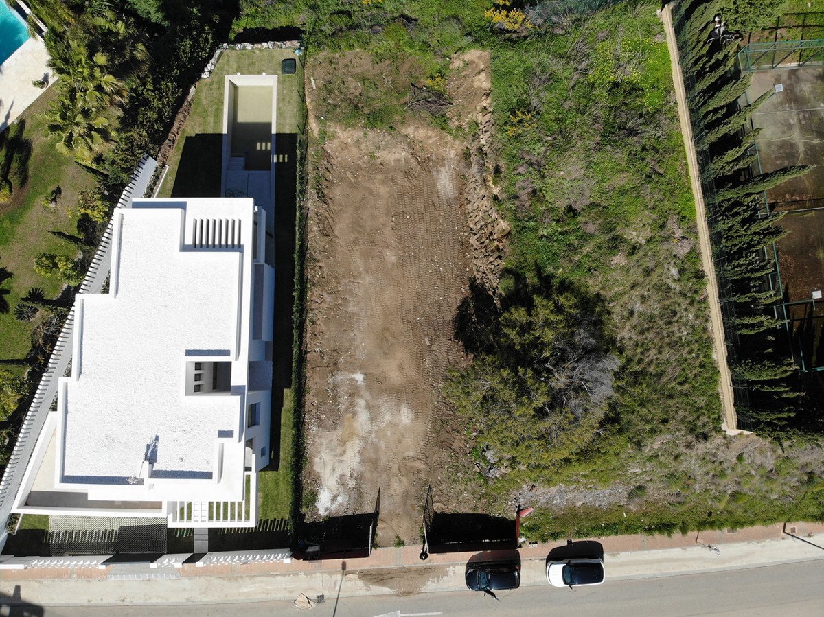 Aerial view of a modern white villa with a pool, adjacent to a cleared construction plot and greenery.