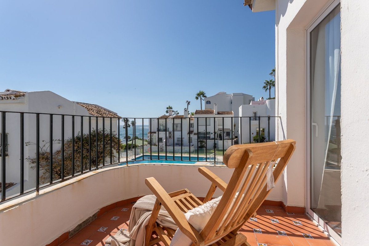 A wooden deck chair on a balcony overlooking a swimming pool and the sea, with white buildings and palm trees.